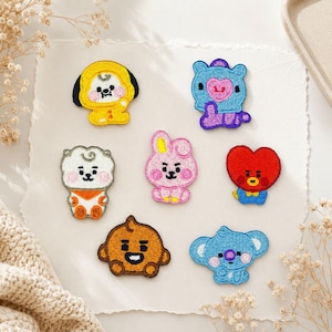 Peut inclure: Un ensemble de sept écussons brodés représentant des personnages de dessins animés colorés. Le lot comprend un chien jaune, une créature bleue avec un cœur rose, un personnage blanc aux accents orange, un lapin rose, un cœur rouge, une figure brune et un koala bleu.