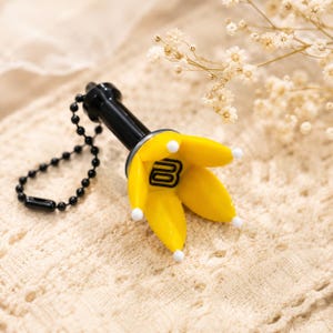 May include: A black keyring featuring a yellow, flower-shaped light stick attachment with five petals. Each petal has a small white tip. The centre of the flower displays a black letter B logo. A black ball chain is attached to the handle.