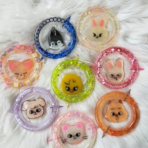 Puede incluir: Una colección de coloridos colgantes de resina con diseños de animales de dibujos animados. Cada colgante tiene un centro transparente con un borde brillante en varios colores, incluyendo azul, rosa, naranja y verde. Los colgantes están dispuestos sobre una superficie blanca y esponjosa.