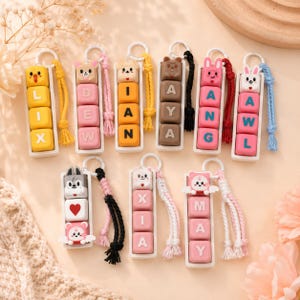 May include: A selection of personalised name keyrings. Each keyring displays a name spelt out on colourful blocks, topped with a cute animal character. The keyrings have a white frame and a braided cord.