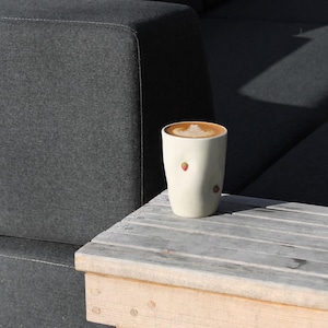 Peut inclure: Un mug en céramique de couleur crème avec une surface irrégulière et bosselée, orné de petits motifs de fraises rouges. Le mug est rempli de café avec un motif de latte art en forme de fougère blanche sur le dessus, contrastant avec le liquide.