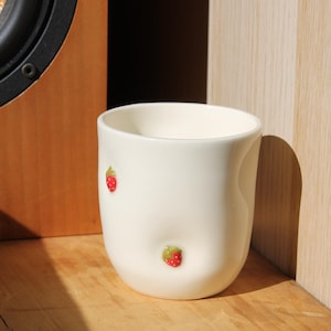 Peut inclure: Une tasse en céramique de couleur crème avec une forme organique légèrement irrégulière. La surface extérieure présente deux petites fraises rouges en relief peintes à la main avec des feuilles vertes, placées à différentes hauteurs.