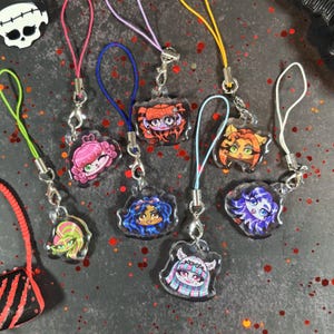 以下が含まれることがあります： 漫画のキャラクターデザインが施された、カラフルなアクリルチャームのコレクション。各チャームは、鮮やかな髪と顔の特徴を持つユニークなキャラクターで、金属製の留め金が付いたストラップに取り付けられています。チャームはさまざまな形とサイズがあります。