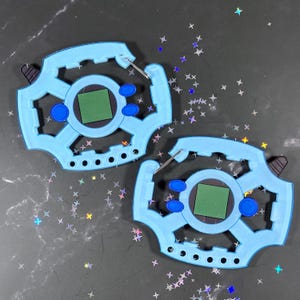 May include: Two light blue plastic digital pet devices with a circular center, a square green screen, and two blue oval buttons on each side. The devices feature a notched outer frame, a small black tab on the side, and a silver metal carabiner clip.