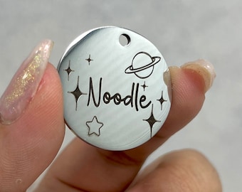 May include: A silver round tag with a planet and stars design. The tag is engraved with the name "Noodle".