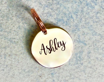 May include: A round, rose gold-colored metal tag with a ring attached. The tag has the name "Ashley" engraved in black script.