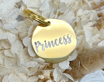 May include: Gold-colored metal pet tag with a round shape and a ring for attaching to a collar. The tag has the word "Princess" engraved on it.