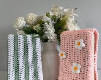 May include: Two crochet book covers. One is striped green and white, the other is pink with white and yellow flower accents. Both covers are handmade and appear to be made of yarn. The covers are standing upright on a white surface.