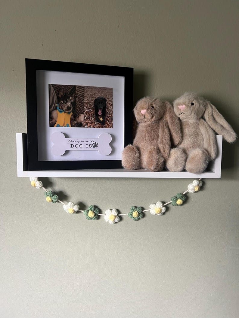 Puede incluir: Un marco de fotos negro que contiene dos fotos de perros y un recorte blanco en forma de hueso con el texto "Home is where the DOG IS" y una huella. Dos conejos de peluche beige se sientan en el estante blanco junto a una guirnalda de flores.
