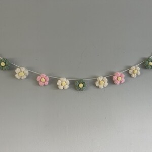 Puede incluir: Una guirnalda con nueve flores de ganchillo ensartadas en un cord&oacute;n blanco fino. Las flores alternan p&eacute;talos de color verde salvia, crema y rosa suave, cada una con un peque&ntilde;o centro amarillo. El conjunto se muestra en forma de arco.
