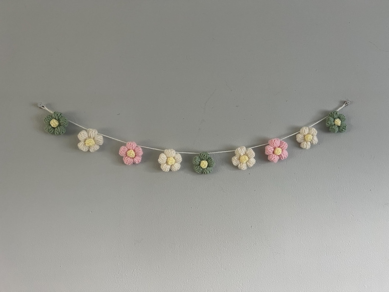 Puede incluir: Una guirnalda con nueve flores de ganchillo ensartadas en un cord&oacute;n blanco fino. Las flores alternan p&eacute;talos de color verde salvia, crema y rosa suave, cada una con un peque&ntilde;o centro amarillo. El conjunto se muestra en forma de arco.