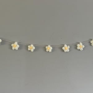 Puede incluir: Una guirnalda con siete flores de ganchillo ensartadas en un cord&oacute;n gris fino. Cada flor tiene cinco p&eacute;talos blancos y un peque&ntilde;o centro amarillo. Las flores est&aacute;n espaciadas uniformemente a lo largo del cord&oacute;n, que tiene lazos en los extremos.