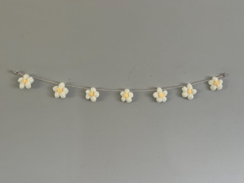 Puede incluir: Una guirnalda con siete flores de ganchillo ensartadas en un cord&oacute;n gris fino. Cada flor tiene cinco p&eacute;talos blancos y un peque&ntilde;o centro amarillo. Las flores est&aacute;n espaciadas uniformemente a lo largo del cord&oacute;n, que tiene lazos en los extremos.
