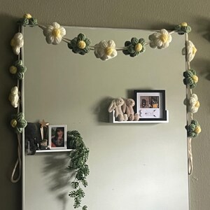 Puede incluir: Una guirnalda decorativa con flores de ganchillo en tonos crema, verde salvia y amarillo, ensartadas en un cord&oacute;n grueso. Las flores est&aacute;n dispuestas en colores y tama&ntilde;os alternos, dise&ntilde;adas para rodear el marco de un espejo rectangular.