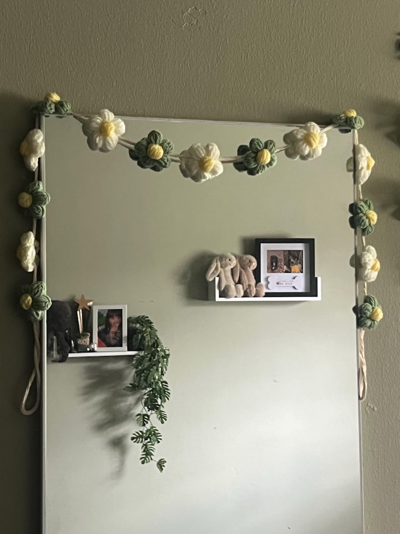 Puede incluir: Una guirnalda decorativa con flores de ganchillo en tonos crema, verde salvia y amarillo, ensartadas en un cord&oacute;n grueso. Las flores est&aacute;n dispuestas en colores y tama&ntilde;os alternos, dise&ntilde;adas para rodear el marco de un espejo rectangular.