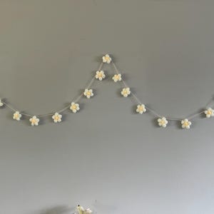 Puede incluir: Una guirnalda decorativa que presenta una serie de peque&ntilde;as flores blancas de ganchillo con centros de color amarillo p&aacute;lido, ensartadas en un cord&oacute;n fino de color claro. Las flores est&aacute;n espaciadas uniformemente a lo largo del hilo.