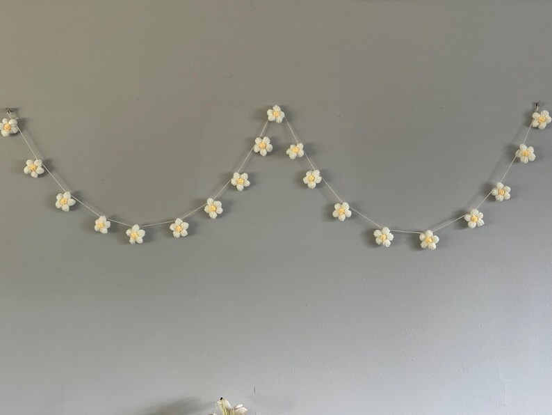 Puede incluir: Una guirnalda decorativa que presenta una serie de peque&ntilde;as flores blancas de ganchillo con centros de color amarillo p&aacute;lido, ensartadas en un cord&oacute;n fino de color claro. Las flores est&aacute;n espaciadas uniformemente a lo largo del hilo.