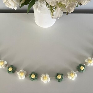 Puede incluir: Una guirnalda de ganchillo con flores alternas blancas y verde salvia con peque&ntilde;os centros amarillos, ensartadas en un cord&oacute;n blanco fino. La tira floral est&aacute; dispuesta en forma curva, mostrando los p&eacute;talos tejidos individualmente.