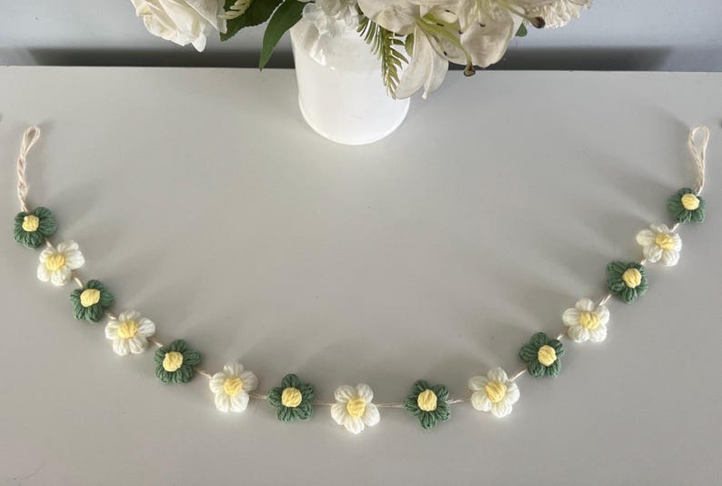 Puede incluir: Una guirnalda de ganchillo con flores alternas blancas y verde salvia con peque&ntilde;os centros amarillos, ensartadas en un cord&oacute;n blanco fino. La tira floral est&aacute; dispuesta en forma curva, mostrando los p&eacute;talos tejidos individualmente.