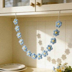 Puede incluir: Una guirnalda decorativa que presenta una serie de flores de ganchillo de color azul claro con peque&ntilde;os centros amarillos, ensartadas en un cord&oacute;n blanco fino. Las flores est&aacute;n dispuestas en forma de U, creando un aspecto suave y texturizado.