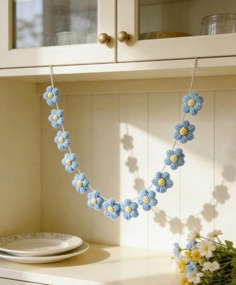Puede incluir: Una guirnalda decorativa que presenta una serie de flores de ganchillo de color azul claro con peque&ntilde;os centros amarillos, ensartadas en un cord&oacute;n blanco fino. Las flores est&aacute;n dispuestas en forma de U, creando un aspecto suave y texturizado.