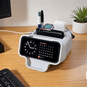 May include: A light gray desk organizer with a digital clock and calendar display. It holds a smartphone, smartwatch, wireless earbuds, and pens. A small potted plant sits nearby. The desk is made of wood.