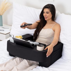 May include: A black bed tray with a built-in phone holder, a tablet stand, and a cup holder. A person is sitting in bed using the tray to hold a remote control, a tablet, a snack, and a cup.