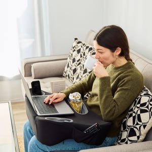 May include: A black laptop desk with built-in cup holders, a phone slot, and a pocket for a remote control. The desk is on a person's lap, with a laptop, phone, and snacks visible. The person is sitting on a gray couch.