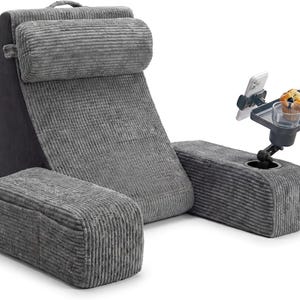 May include: Gray corduroy reading pillow with a headrest, armrests, and a footrest. Includes a side table with a phone holder, cup holder, and a small tray holding a muffin. The pillow has a handle for easy carrying.