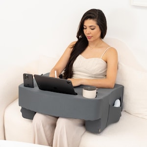 May include: A gray laptop desk with a built-in cup holder and phone slot. The desk is designed to be used on a couch or bed. A person is sitting on a couch and using the desk to work on a laptop.