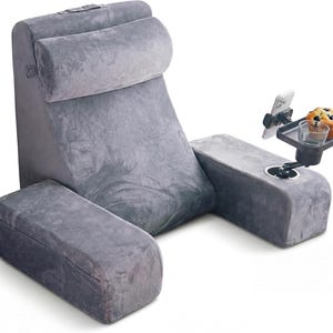 May include: A grey reading pillow with a headrest, armrests, and a footrest. It features a side tray with a cup holder, a phone holder, and a muffin. The pillow is designed for comfort and relaxation, ideal for reading.