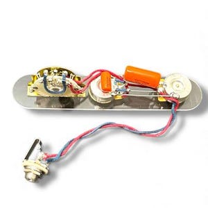 May include: Guitar control plate with electronic components. The plate is silver with three potentiometers, orange capacitors, and wiring. A chrome jack is connected with red and blue wires. The components are arranged on a rectangular plate.