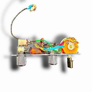 May include: Close-up of a guitar wiring harness with various colored wires, orange capacitor, and a yellow switch. The harness is mounted on a silver metal plate with three silver knobs and a gold-colored output jack.