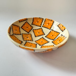 Puede incluir: Cuenco de cerámica ovalado con interior y exterior color crema. El cuenco está decorado con un patrón repetido de formas de galletas saladas naranjas delineadas en negro. Las galletas saladas tienen pequeños puntos negros en el centro.