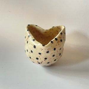 May include: A small, white ceramic bowl with a speckled brown glaze and black polka dots. The bowl is shaped like a heart and has a wide opening.