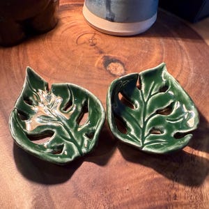 May include: Two ceramic leaf-shaped dishes with a glossy green glaze. The dishes have a monstera leaf design with cut-out sections. The dishes are on a wooden surface.
