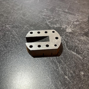 May include: A small, metallic, U-shaped object with multiple circular holes. The object has a rectangular base and a slot cut out of the center. The metal has a slightly worn appearance.