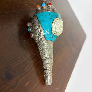 May include: A decorative silver-toned object with turquoise and colorful stone accents. The object has an ornate design with a detailed, textured pattern. The top features turquoise stone and is adorned with red, blue, and orange stones.