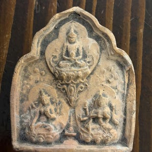 May include: A weathered, rectangular, tan-colored religious amulet. The amulet features three figures: a central seated figure and two flanking figures. The figures are in relief, with intricate details. The background is a darker, contrasting color.