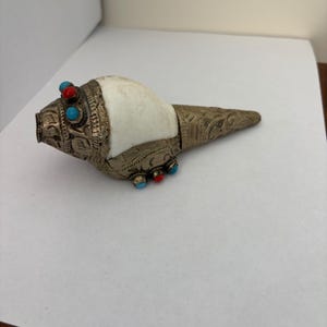 Puede incluir: Una concha de caracola ornamentada, de color plateado, con detalles turquesa y rojos. La concha tiene una superficie blanca y lisa y está decorada con intrincados trabajos en metal. El diseño presenta un patrón detallado y es probablemente un objeto decorativo.