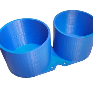 Puede incluir: Un portavasos doble de plástico azul impreso en 3D con dos vasos cilíndricos. Los vasos están conectados por una base con agujeros.