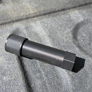 Puede incluir: Un objeto cil&iacute;ndrico de metal negro con un extremo texturizado y estriado y una base hexagonal. El objeto est&aacute; sobre una superficie gris texturizada, proyectando una sombra.