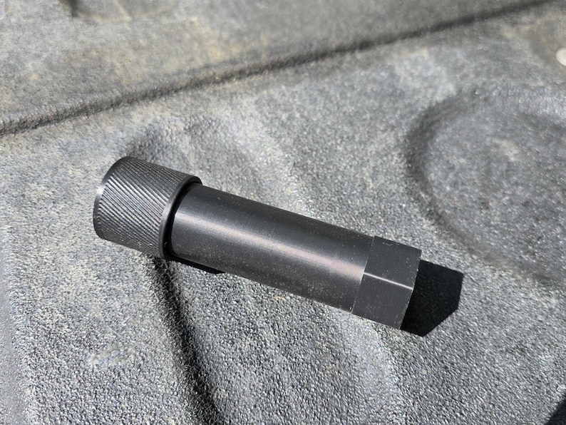 Puede incluir: Un objeto cil&iacute;ndrico de metal negro con un extremo texturizado y estriado y una base hexagonal. El objeto est&aacute; sobre una superficie gris texturizada, proyectando una sombra.