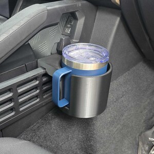 Peut inclure: Un mug de voyage en acier inoxydable avec une poignée bleue et un couvercle transparent est placé dans un porte-gobelet noir. Le porte-gobelet est monté dans la console d'un véhicule. Le mug a une bande argentée en haut et une bande bleue en bas.