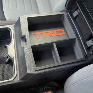 以下が含まれることがあります： 赤色の「TRD」ロゴが付いた黒い車のコンソールオーガナイザー。 オーガナイザーには2つのコンパートメントがあり、車両のセンターコンソール内に収まります。 内装はグレーで、周囲のコンソールは黒です。