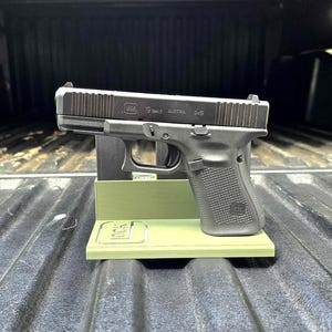 Könnte beinhalten: Eine schwarze Glock 19 Gen 5 Pistole steht auf einem hellgrünen Ständer. Die Pistole ist mit "Glock 19 Gen 5 AUSTRIA 9x19" gekennzeichnet. Der Ständer hat das Glock-Logo.