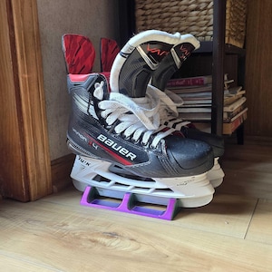 May include: A pair of black and white ice skates with red accents. The skates are on a purple skate rack. The skates have the word "Bauer" printed on the side.