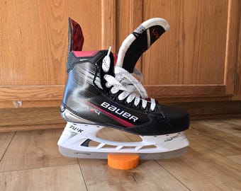 May include: Black and red ice hockey skate with white laces and a white Bauer logo. The skate is on a wooden floor with a small orange block underneath.