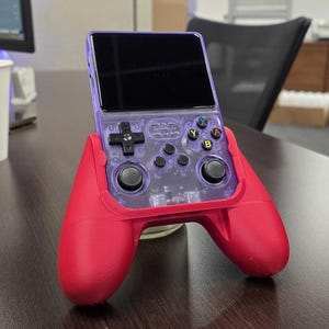 May include: A handheld gaming console with a transparent purple shell and a red controller grip. The console features a black screen, directional pad, and colored action buttons. The device is resting on a wooden surface.