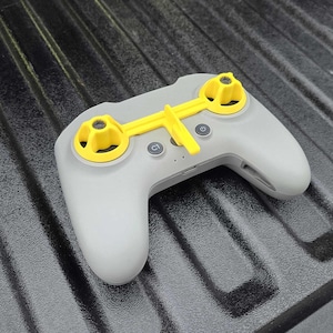 May include: A gray remote control with yellow accents. The remote has two yellow control wheels and a yellow bar connecting them. It features buttons labeled "C1" and a power button. The device is designed for drone operation.